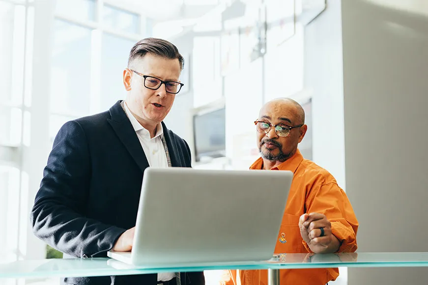 Two office workers at a standing desk are discussing a the issue they see on the laptop before them